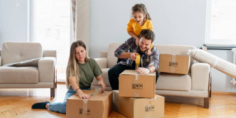 Happy family parents and children tenants renters new home owners playing on moving day unpacking boxes, cute little child daughter jump on her father's neck